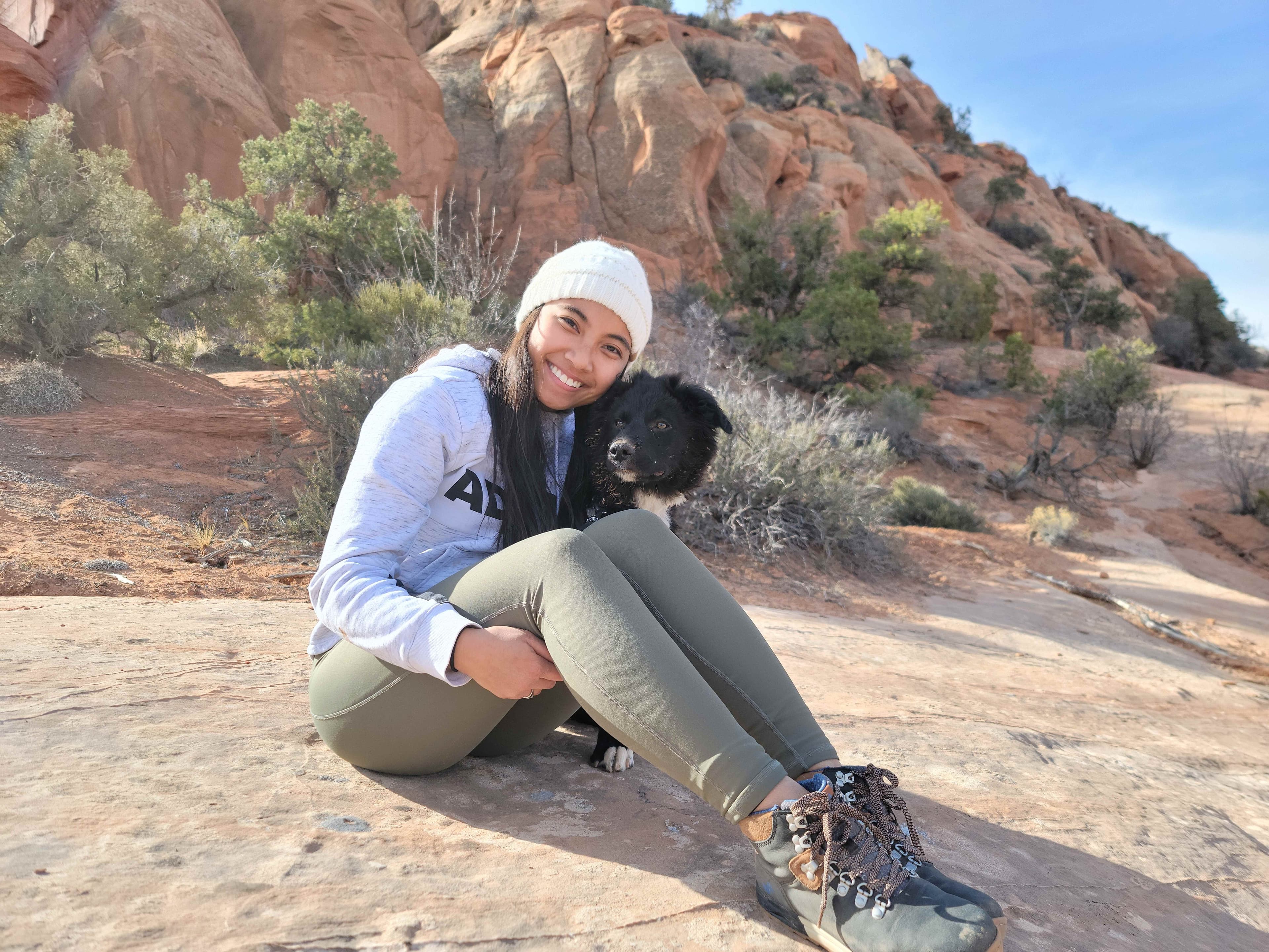 Katrina sitting on red sandstone in the Utah desert with her black dog, wearing a white beanie and grey hoodie, with red rock formations in the background