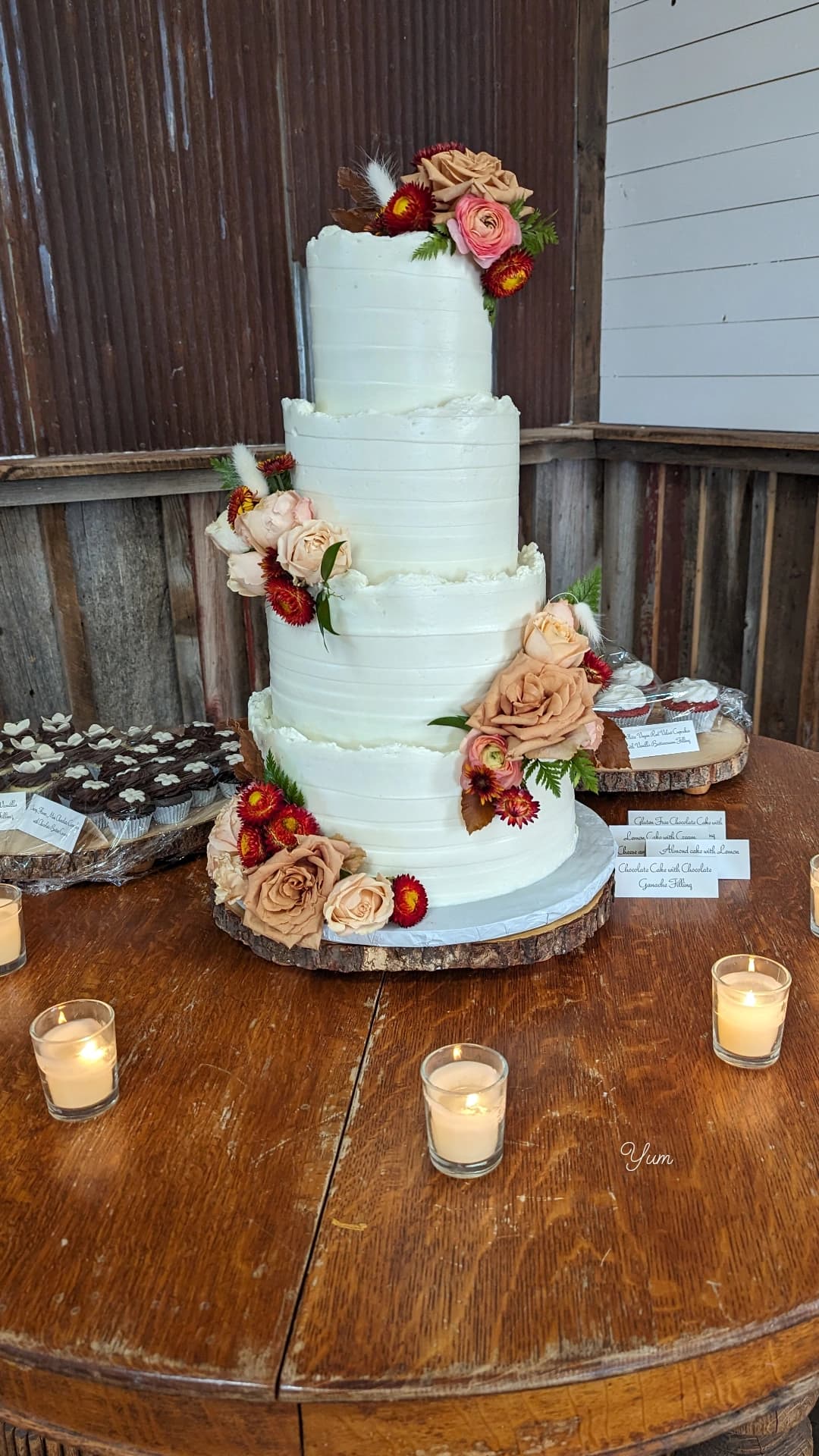 Four-tier white textured wedding cake with cascading terracotta, blush, and peach blooms on a wood slice stand with votive candles