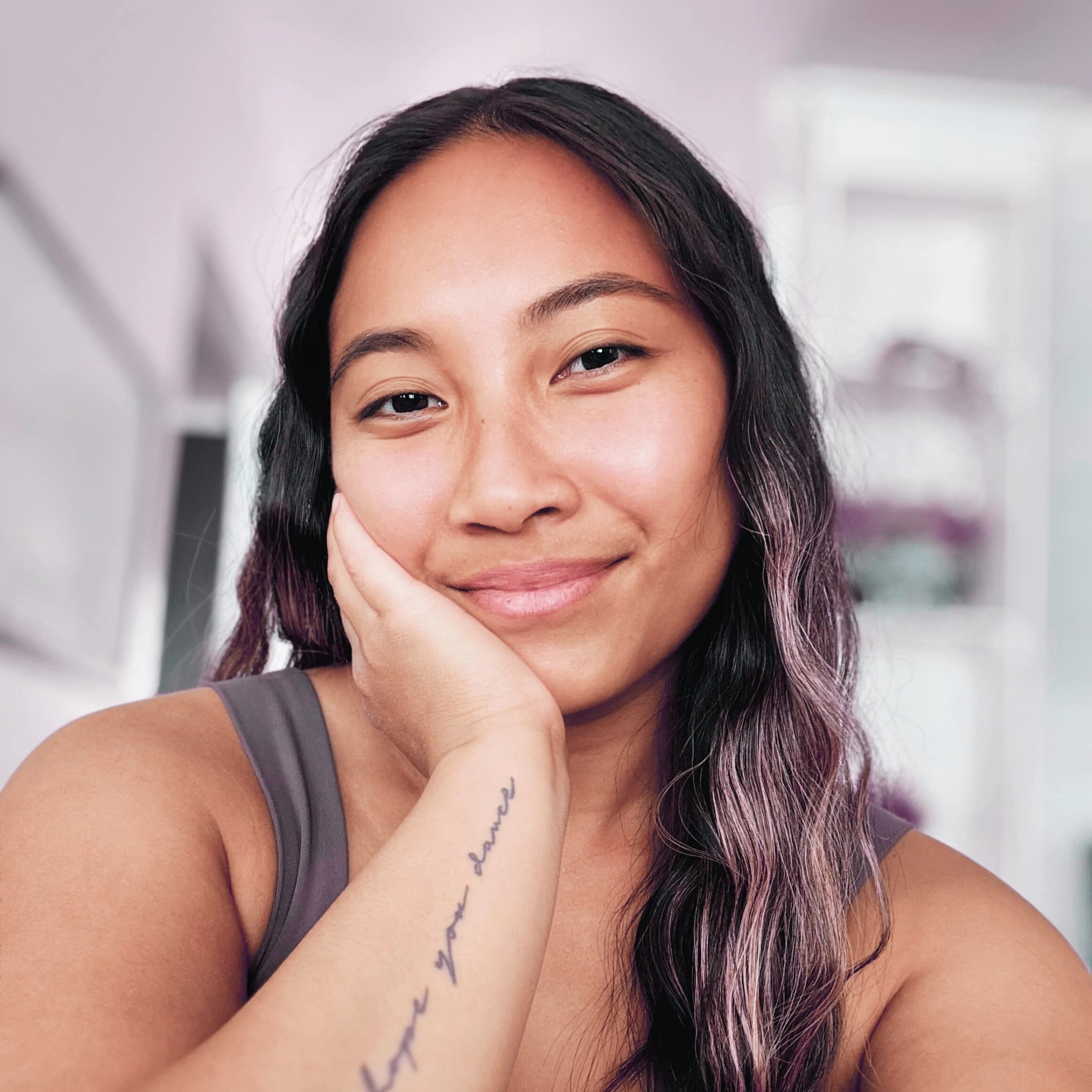 Katrina Mae, a Filipino-American woman with long dark wavy hair, resting her chin on her hand with a soft smile, script tattoo visible on her forearm