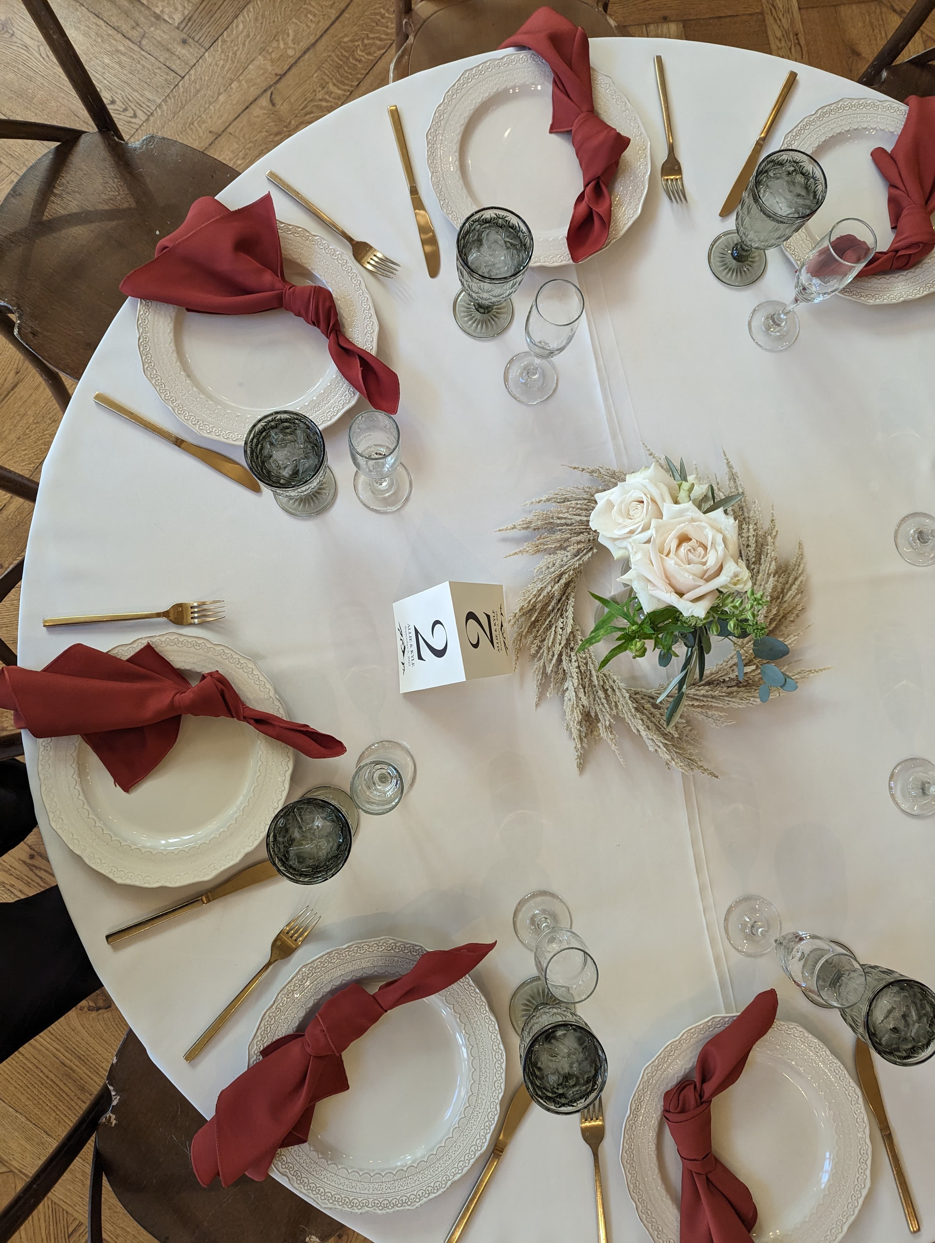 Top-down view of round reception table with white plates, burgundy napkins, gold flatware, and blush roses with pampas grass centerpiece
