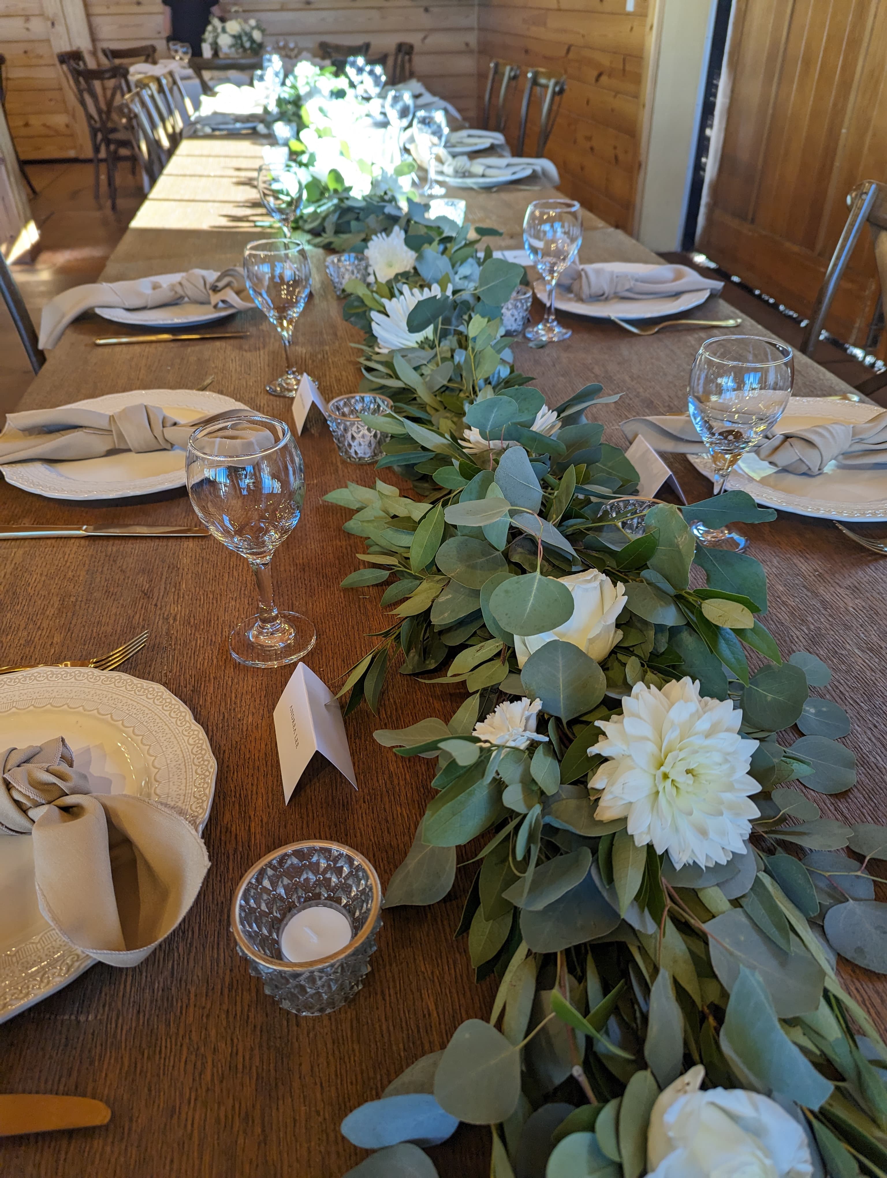 Long farm table with eucalyptus garland, white dahlias and roses, taupe napkins, and clear glassware in a wood-paneled barn